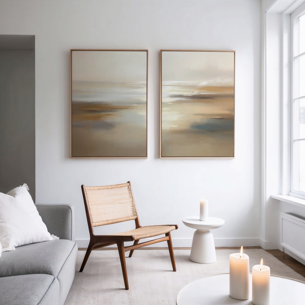 A minimalist living room featuring a woven chair, round white side table, three lit candles, light gray sofa with white pillows, and the "Sunlit Waters - Set Of 2" wall art on a white wall near a large sunlit window.