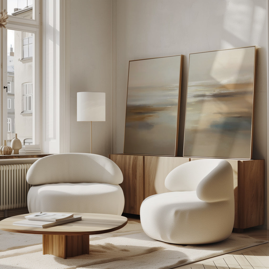A bright, modern living room features the "Sunlit Waters - Set Of 2" tranquil artwork displayed on a low wood sideboard. Two white curved armchairs and a round wooden coffee table complete the calm, minimalist space bathed in sunlight from tall windows.