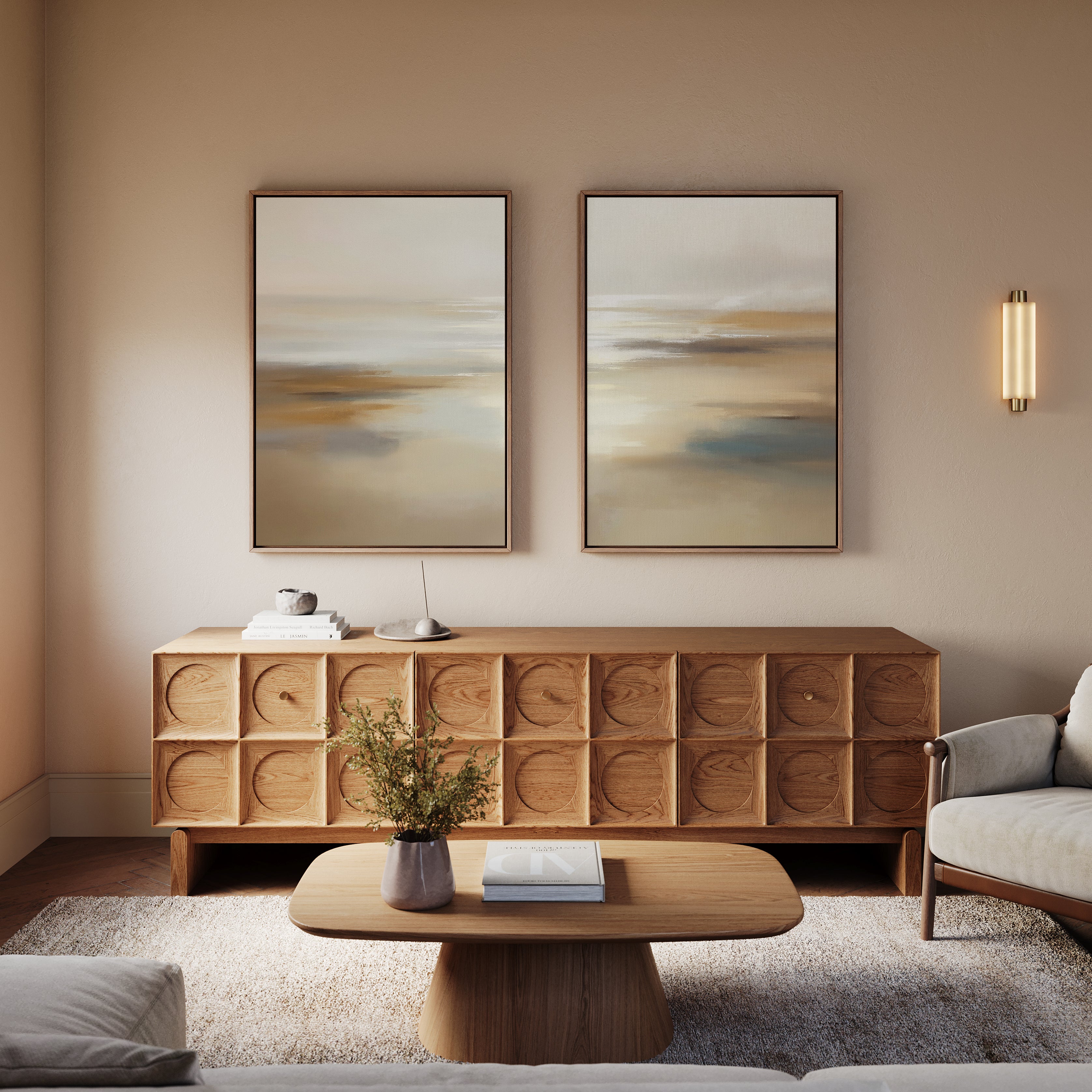 A cozy living room features a wooden sideboard topped by Sunlit Waters - Set Of 2 wall art, a modern wall sconce, beige sofa, and wooden coffee table with a book and vase of greenery for a minimalist, neutral-toned decor.