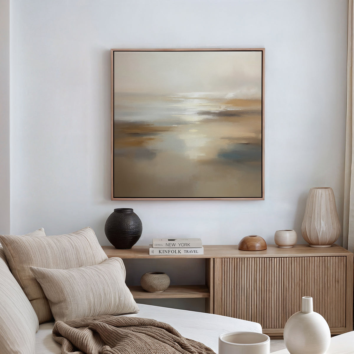 A minimalist living room features a beige sofa, neutral pillows, wooden sideboard, ceramic vases, and stacked books. Above the sofa, the Sunlit Waters Canvas Art - Square adds elegance with its soft brown and gray tones.