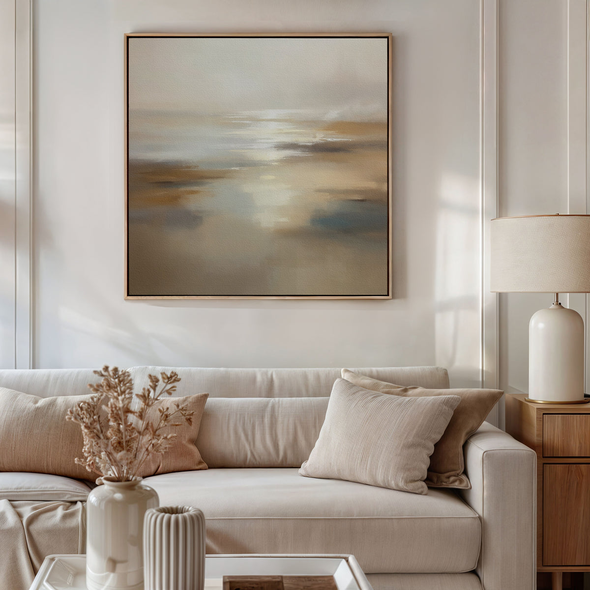 A modern living room with a white sofa, beige cushions, and a side table with a lamp features the Sunlit Waters Canvas Art - Square above. A vase with dried flowers rests on the coffee table underneath.