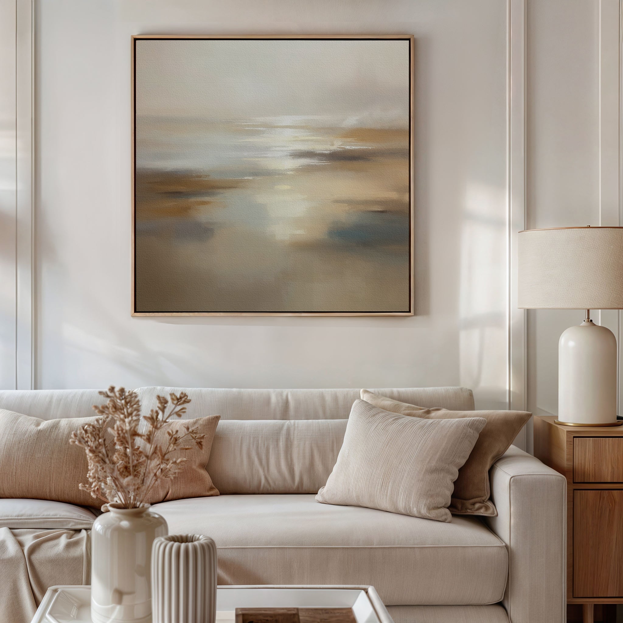 A modern living room with a white sofa, beige cushions, and a side table with a lamp features the Sunlit Waters Canvas Art - Square above. A vase with dried flowers rests on the coffee table underneath.
