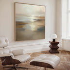 A minimal living room features the Sunlit Waters Canvas Art - Square as a striking focal point, paired with a white leather lounge chair and ottoman, a geometric dark wood side table, and a small white lamp on herringbone wood flooring.