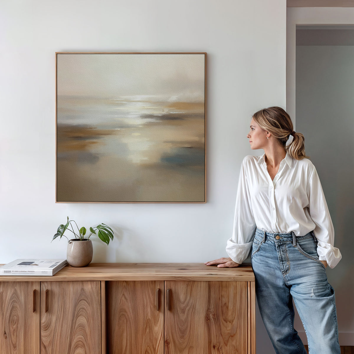 A woman in a white blouse and jeans stands next to a wooden sideboard, admiring the Sunlit Waters Canvas Art - Square, a beige and cream landscape print on the white wall. A small plant and books sit atop the sideboard.