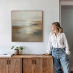 A woman in a white blouse and jeans stands next to a wooden sideboard, admiring the Sunlit Waters Canvas Art - Square, a beige and cream landscape print on the white wall. A small plant and books sit atop the sideboard.