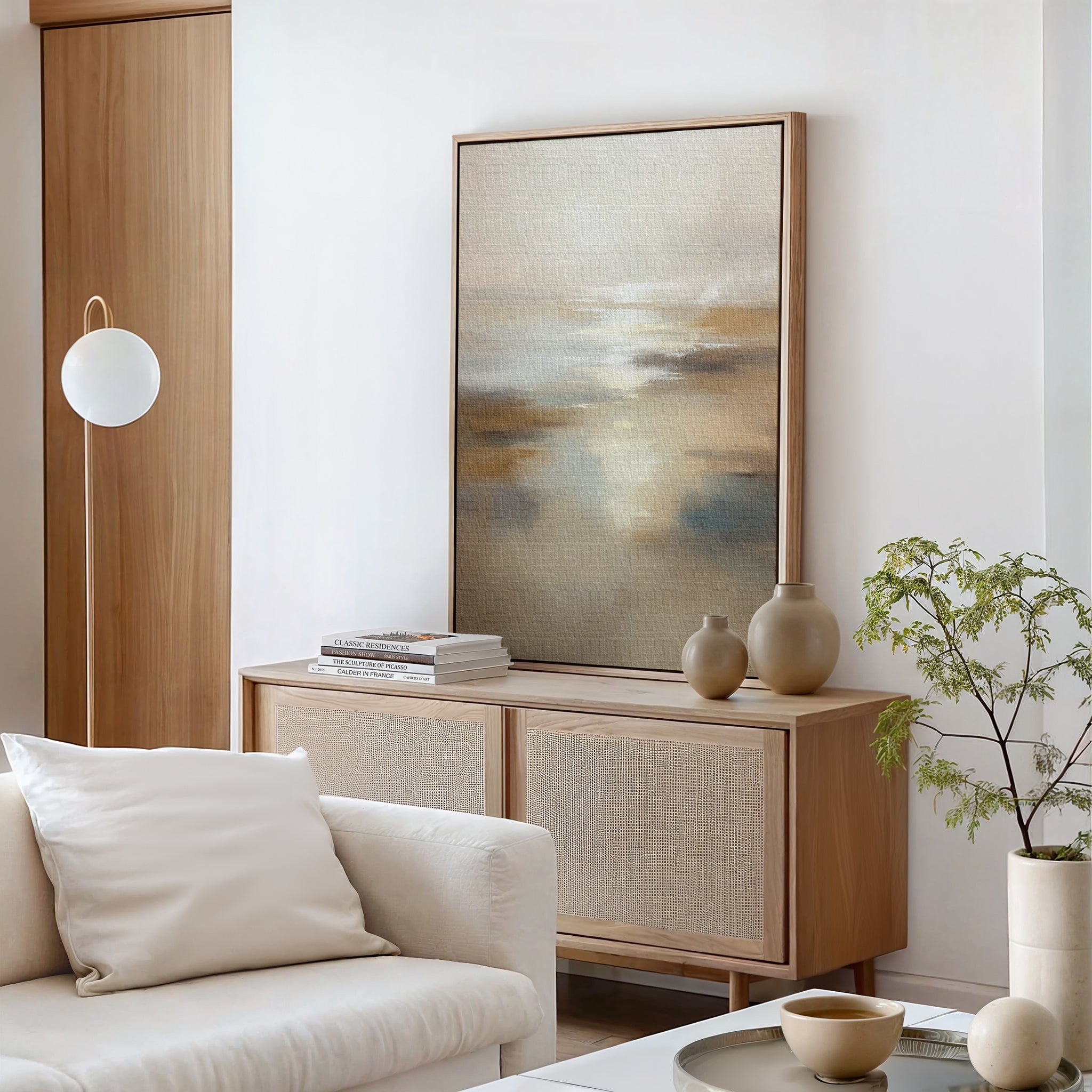 A modern living room with a light wood sideboard, Sunlit Waters Canvas Art - Vertical, beige vases, a small green plant, stacked books, a white sofa with a pillow, and a round pendant light.