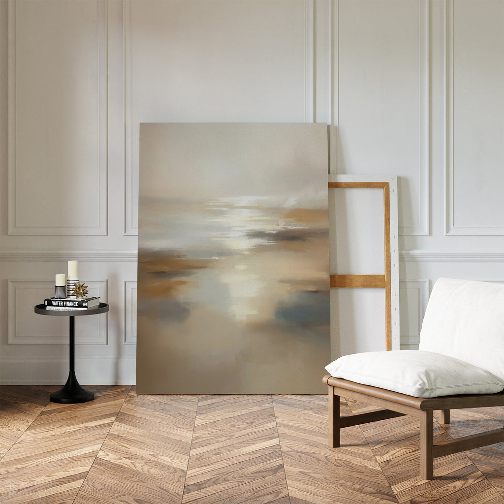 The Sunlit Waters Canvas Art - Vertical, featuring soft muted colors, leans against a white paneled wall beside an empty frame. Nearby are a cushioned chair and small round table with candles and books on a herringbone wood floor.