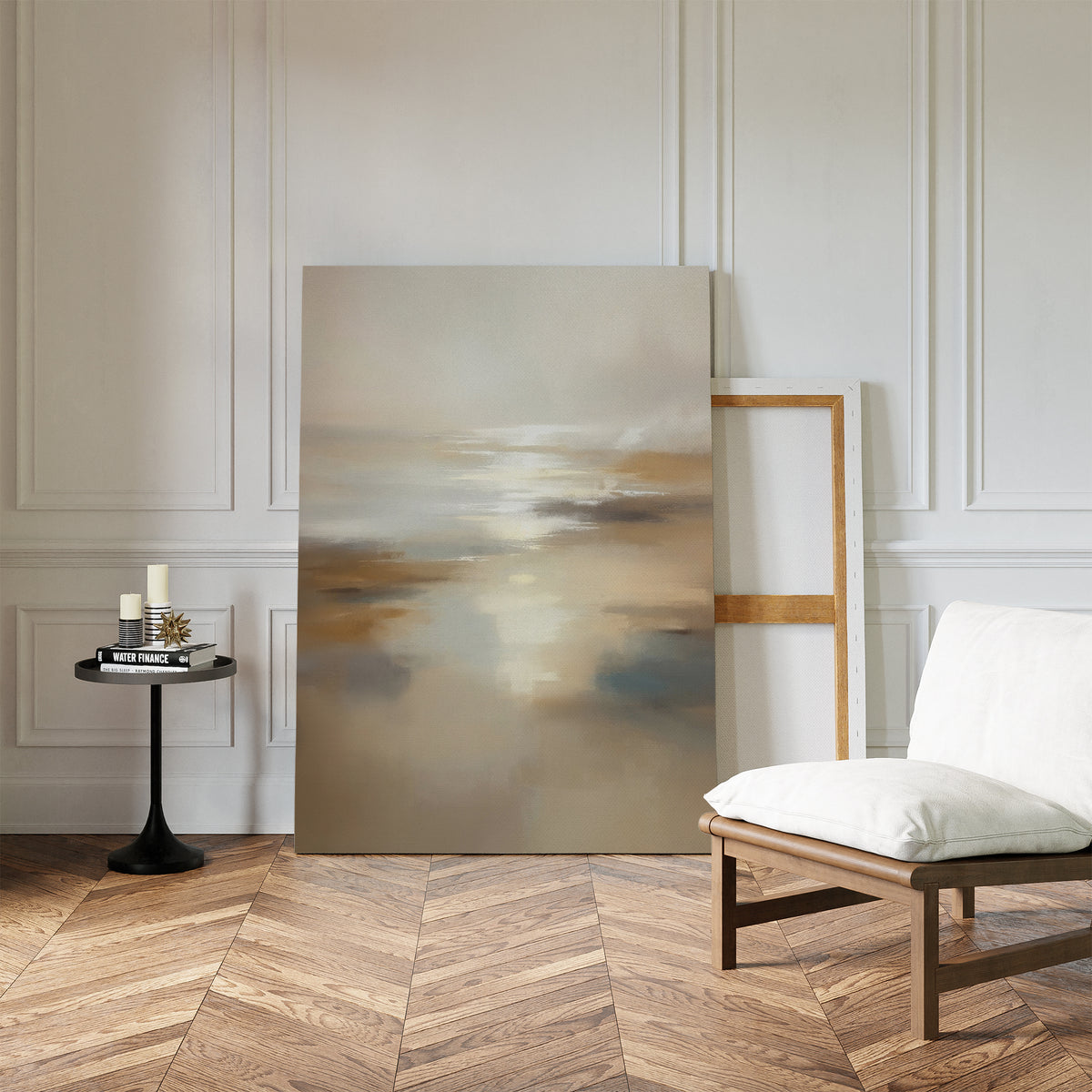 The Sunlit Waters Canvas Art - Vertical, featuring soft muted colors, leans against a white paneled wall beside an empty frame. Nearby are a cushioned chair and small round table with candles and books on a herringbone wood floor.
