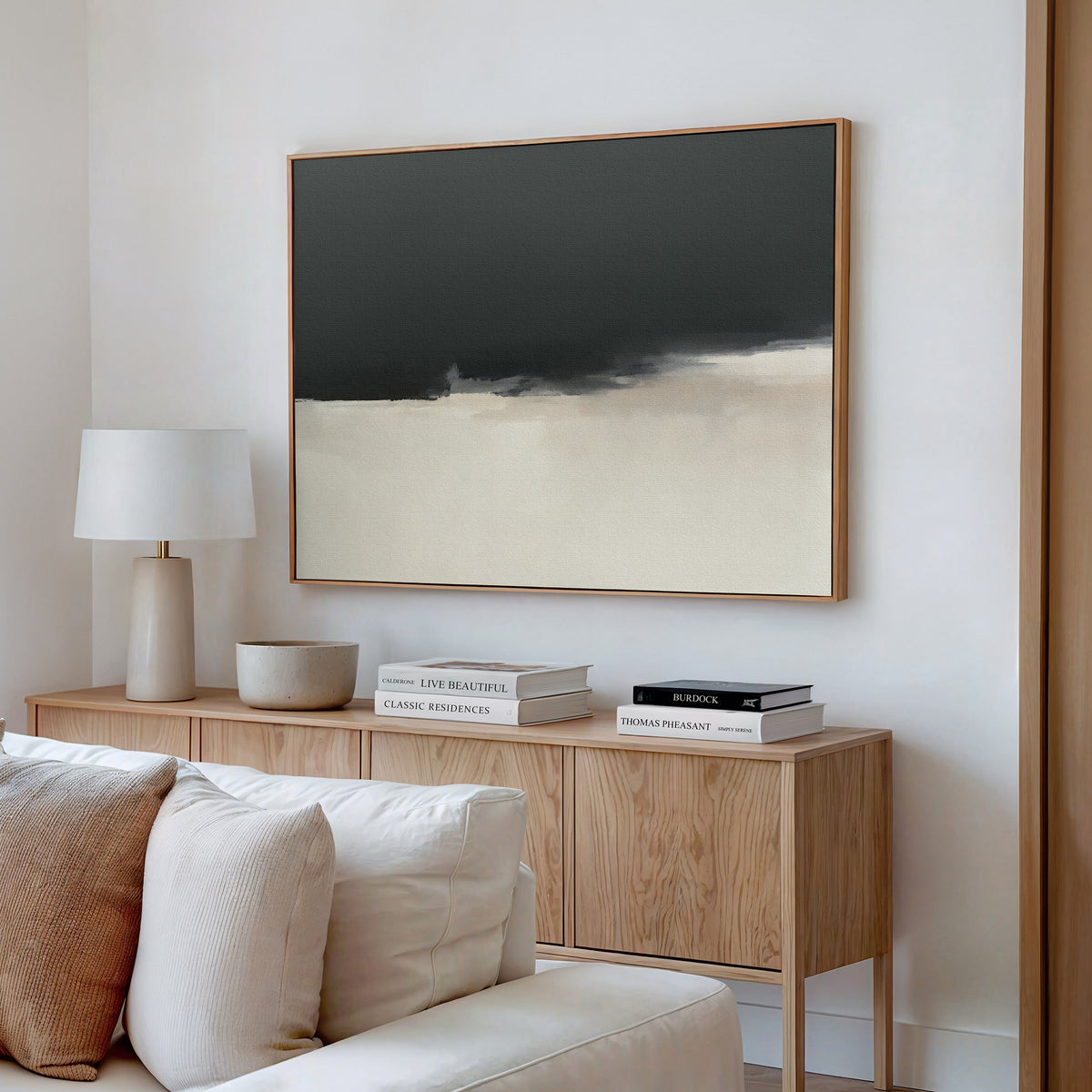 A minimalist living room features a light wood sideboard, a white lamp, stacked books, and the Tempest Brewing Canvas Art - Horizontal—an abstract painting in black, gray, and beige—hung on a white wall above a beige sofa.