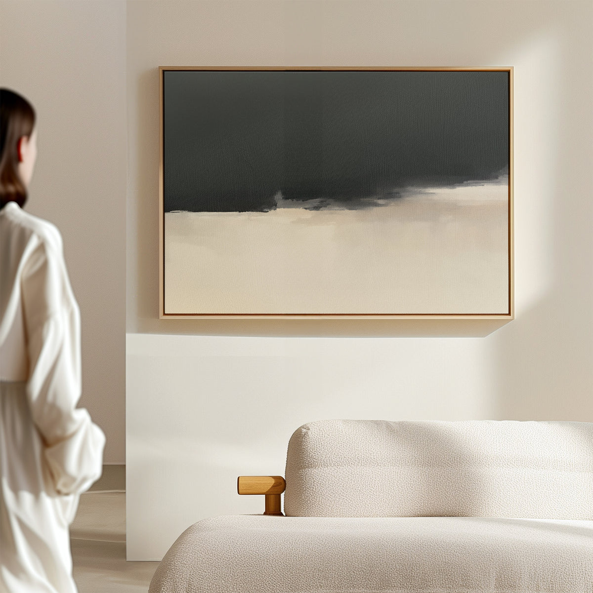 A woman in a white blouse stands in a modern, minimal living room, admiring the Tempest Brewing Canvas Art - Horizontal, a large abstract painting with dark gray and beige hues, hanging above a light-colored sofa.
