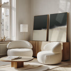 A modern living room features the Tempest Brewing - Set Of 2 rounded white armchairs, a low wooden coffee table, and striking wall art atop a sideboard beneath large windows flooding the space with natural light for an atmospheric vibe.