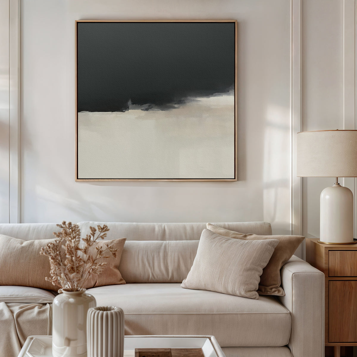 A modern living room with a light beige sofa, neutral pillows, a ceramic table lamp, dried flowers in a vase, and dramatic wall art—the Tempest Brewing Canvas Art - Square in black, white, and beige tones—on the wall.