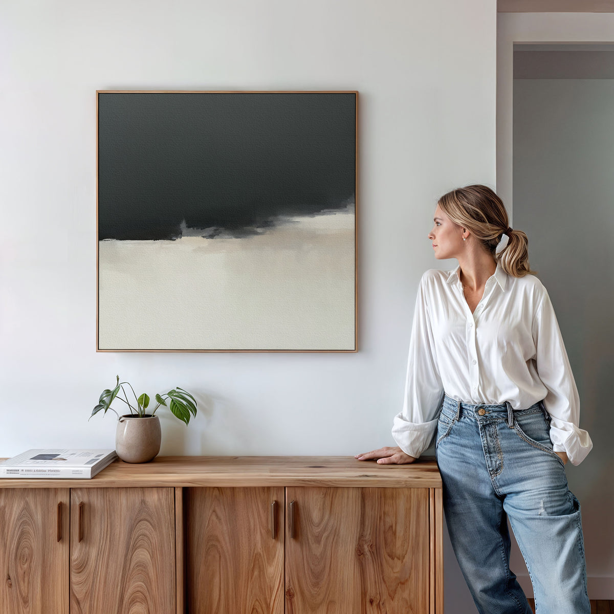 A woman in a white blouse and jeans stands by a wooden sideboard, gazing at the Tempest Brewing Canvas Art - Square. A potted plant and books sit on the sideboard beneath this bold artwork on the white wall.
