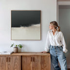 A woman in a white blouse and jeans stands by a wooden sideboard, gazing at the Tempest Brewing Canvas Art - Square. A potted plant and books sit on the sideboard beneath this bold artwork on the white wall.
