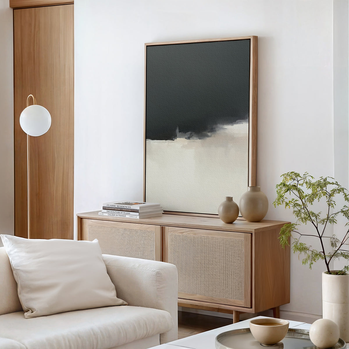 A modern living room with a light wood sideboard, Tempest Brewing Canvas Art - Vertical on the wall, neutral vases, stacked books, a white sofa with cushions, a potted plant, and a round white floor lamp. The decor is minimalist and contemporary.
