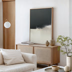 A modern living room with a light wood sideboard, Tempest Brewing Canvas Art - Vertical on the wall, neutral vases, stacked books, a white sofa with cushions, a potted plant, and a round white floor lamp. The decor is minimalist and contemporary.