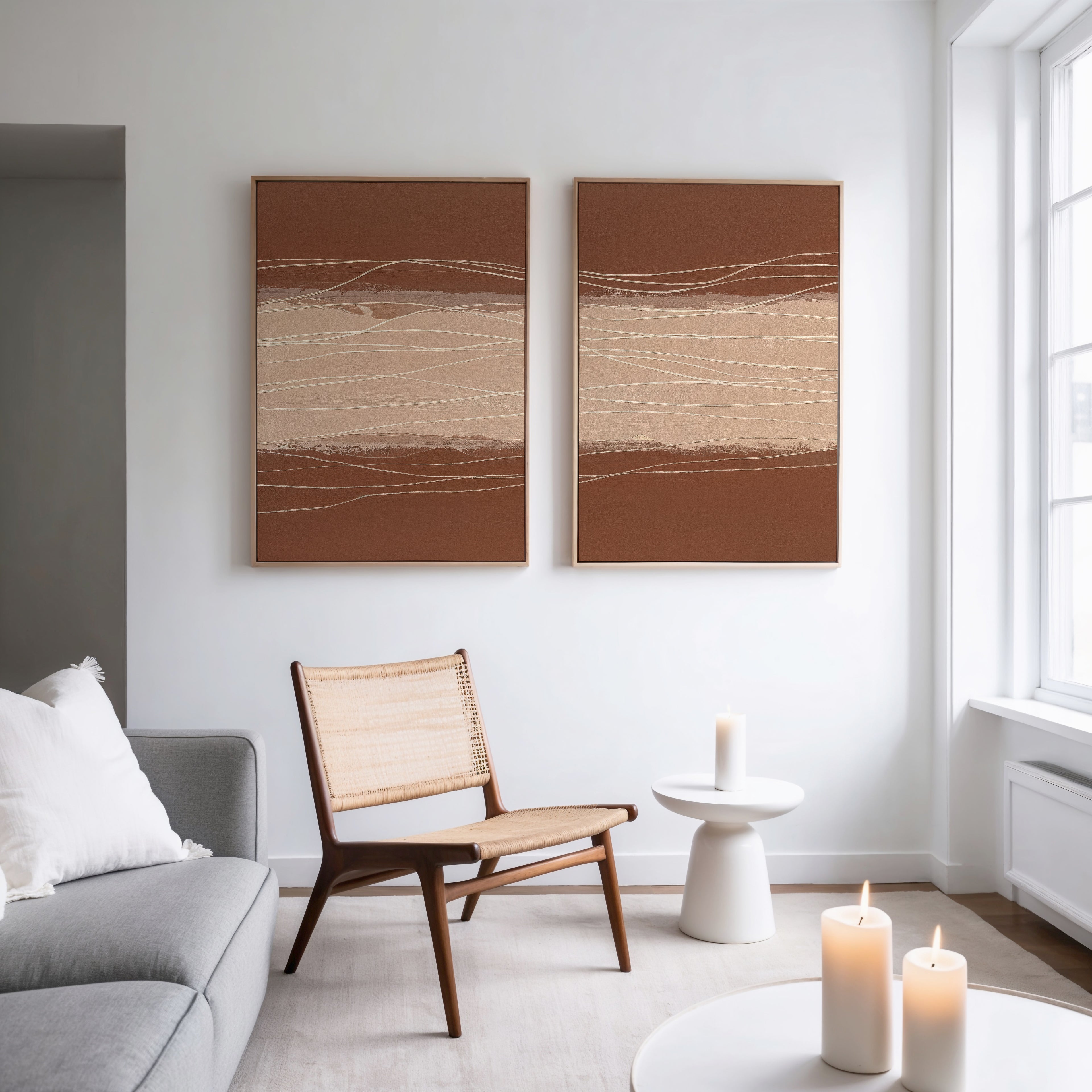 A minimal living room features a gray sofa, wicker chair, small white side table with a candle, and large windows. Modern elegance shines with the Terracotta Currents - Set Of 2 wall art and three glowing candles in the foreground.