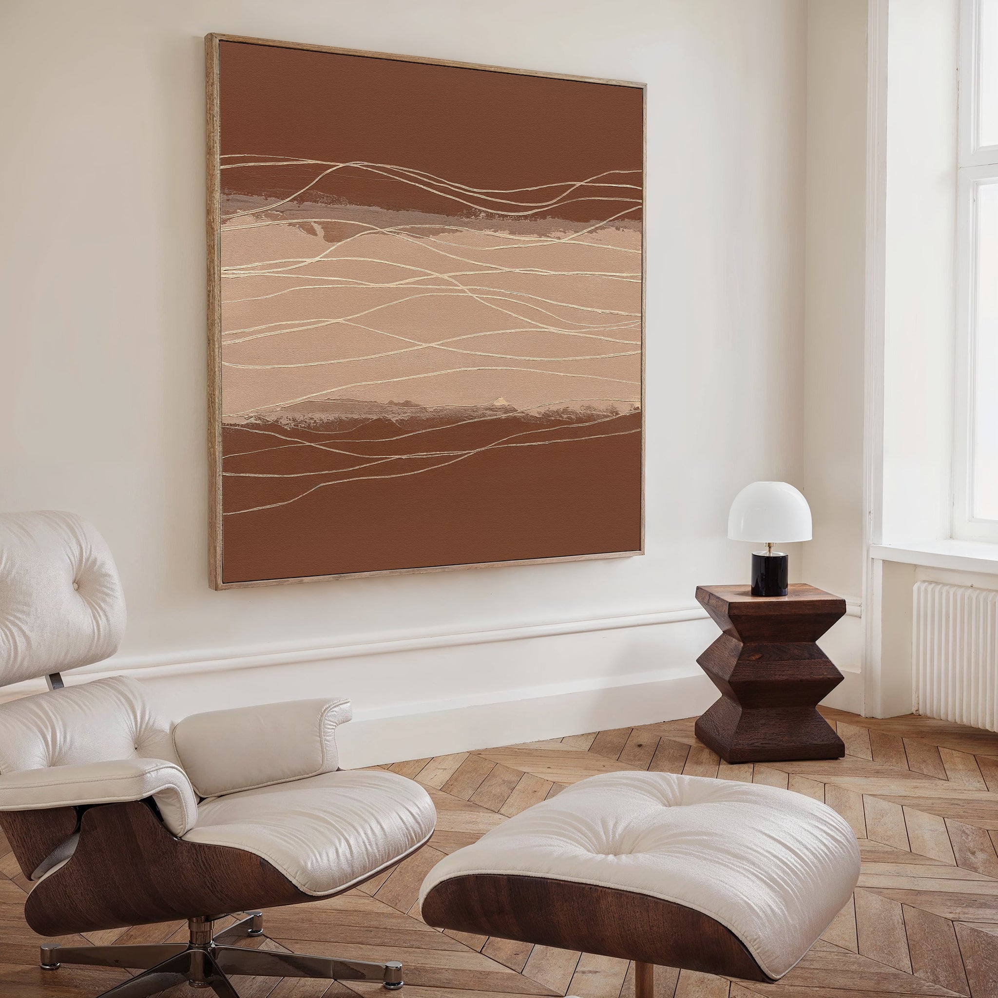 A modern living room with a white leather lounge chair and ottoman, a geometric wooden side table with a white lamp, and the Terracotta Currents Canvas Art - Square in brown and beige tones, adding Nordic Park-inspired style to the space.
