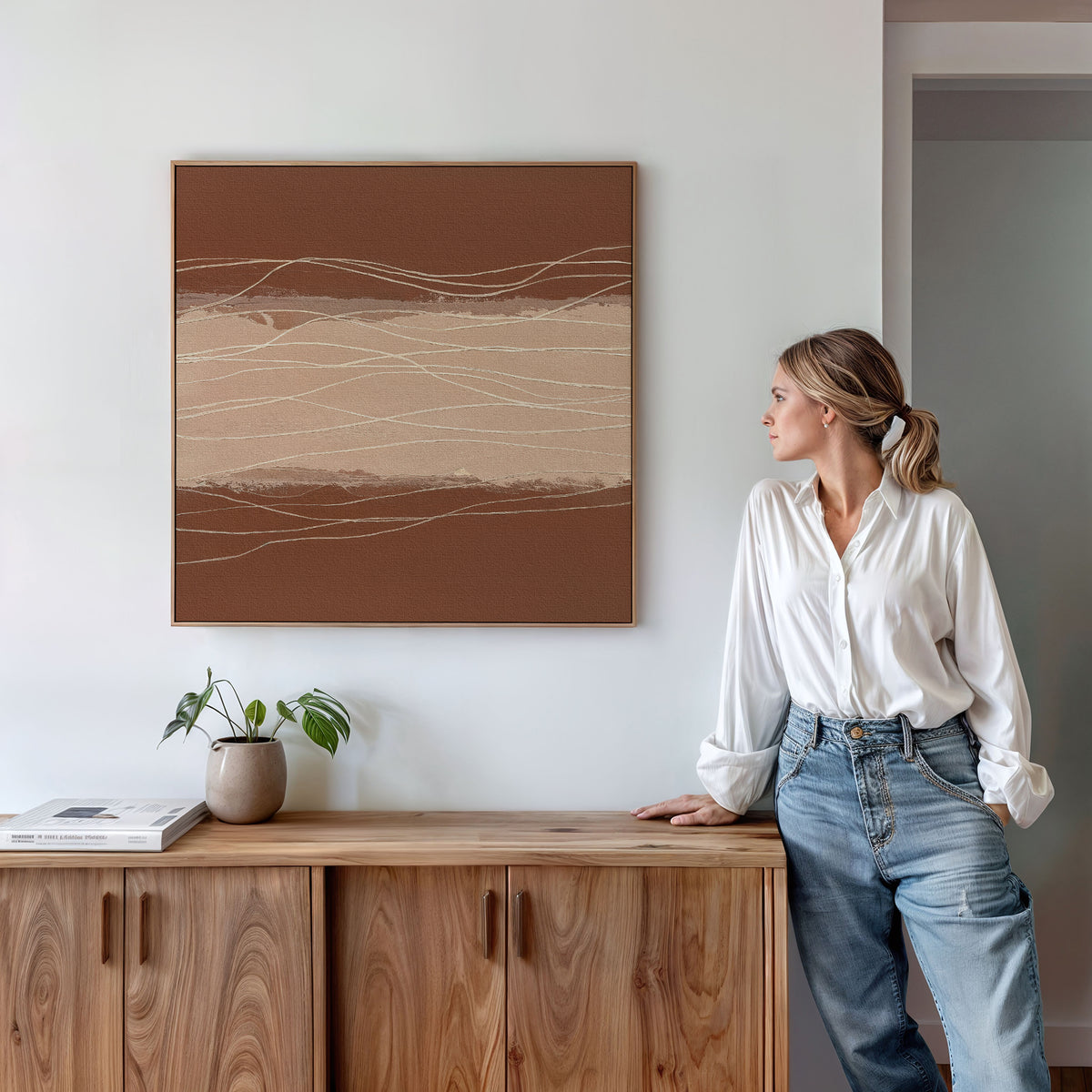 A woman in a white blouse and jeans leans on a wooden cabinet, admiring the Terracotta Currents Canvas Art - Square in earthy tones. A small plant and book rest on the cabinet, enhancing the Nordic Park-inspired decor.