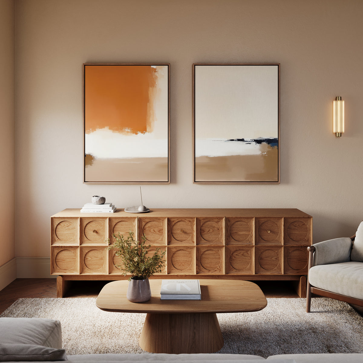A modern living room with a wooden sideboard, Terracotta Edge - Set Of 2 wall art in burnt orange, a round coffee table holding a plant and book, a light gray armchair, soft beige walls, and a wall sconce.