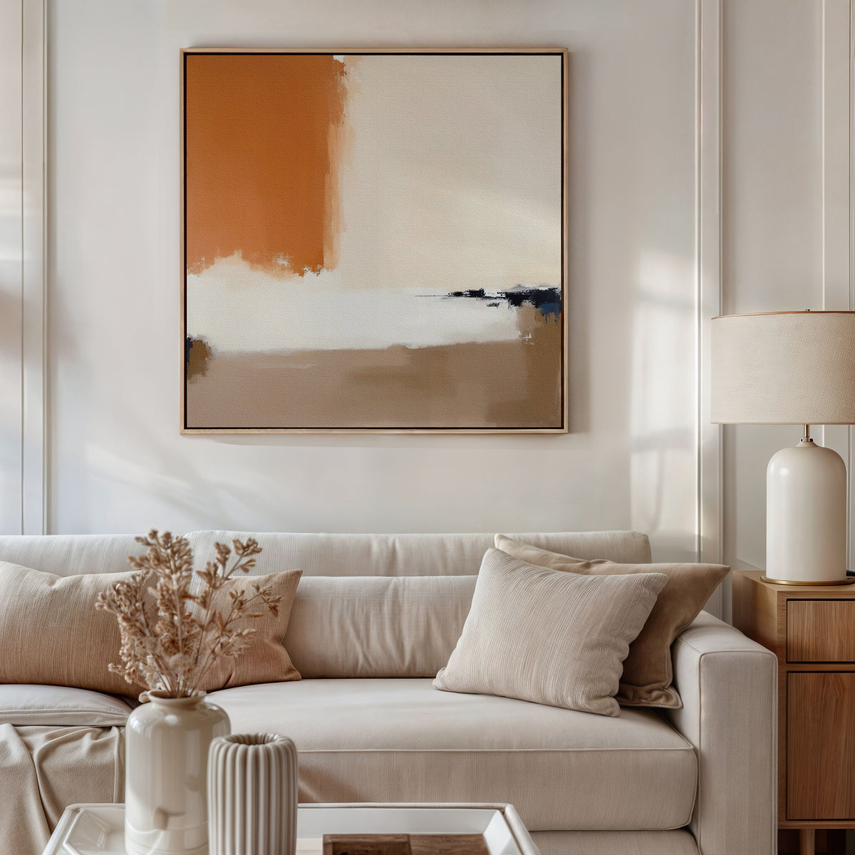 A modern living room with a beige sofa, neutral cushions, and a side table with a ceramic lamp features the Terracotta Edge Canvas Art - Square above the sofa, adding contemporary style in burnt orange, white, and beige tones.