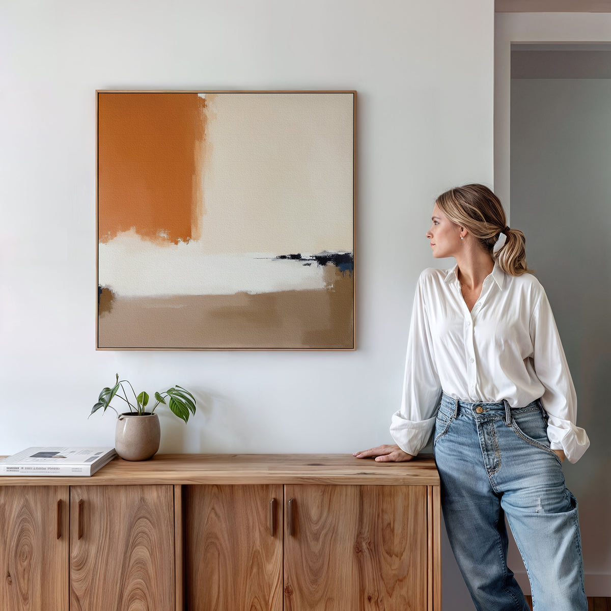 A woman in a white blouse and jeans leans against a wooden cabinet, gazing at the Terracotta Edge Canvas Art - Square above her—a modern abstract print in orange, beige, and white that perfectly accents the living room decor. A potted plant and books rest on the cabinet.