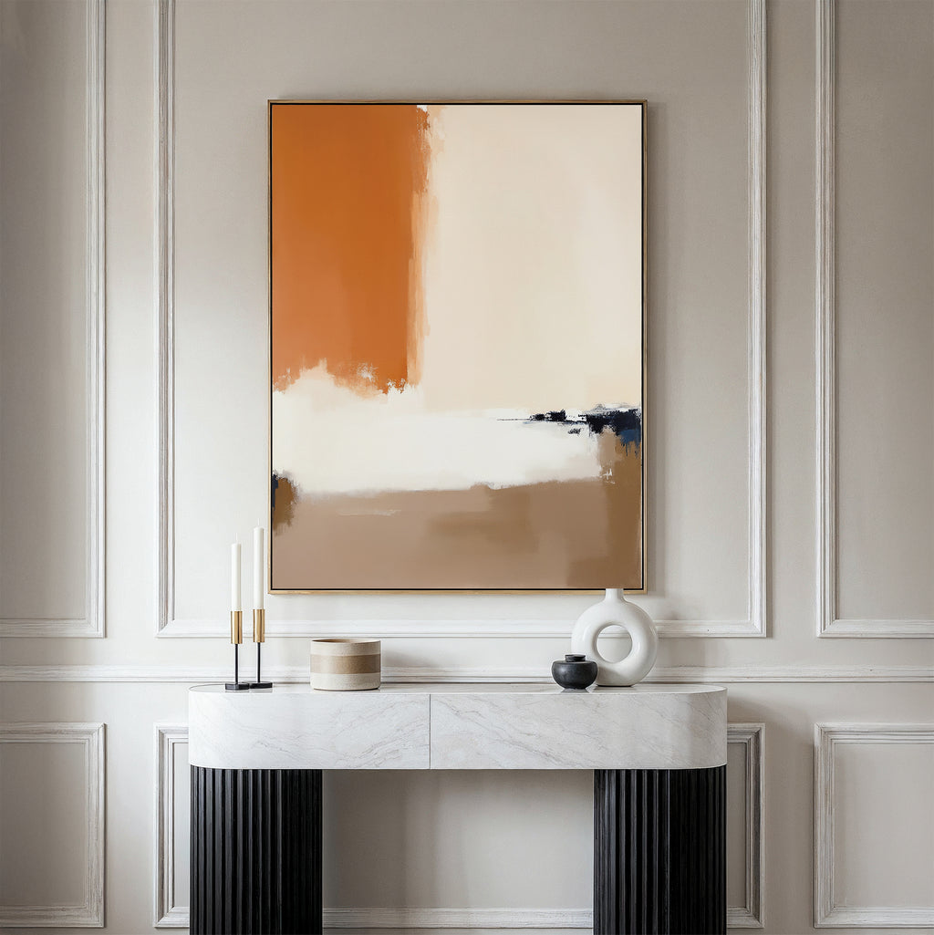 A minimalist console table with a white marble top and black legs stands against a light paneled wall, adorned with candles and decor. Above it hangs the Terracotta Edge Canvas Art - Vertical, featuring beige, black, white, and burnt orange hues.