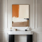 A minimalist console table with a white marble top and black legs stands against a light paneled wall, adorned with candles and decor. Above it hangs the Terracotta Edge Canvas Art - Vertical, featuring beige, black, white, and burnt orange hues.