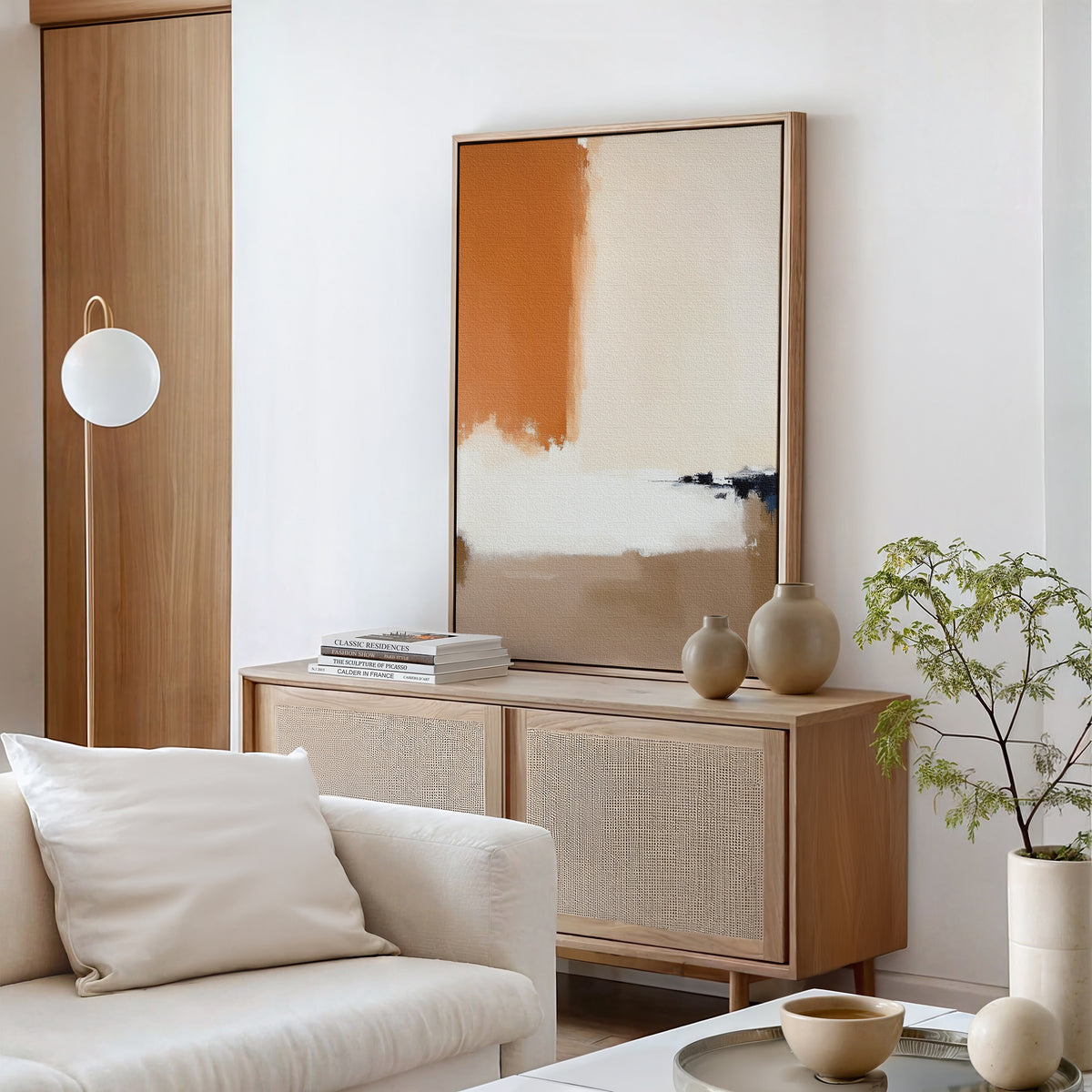 A modern, minimalist living room features a light wood sideboard, neutral ceramics, a potted plant, and a white cushioned sofa. Soft beige and tan decor is accented by the Terracotta Edge Canvas Art - Vertical and contemporary abstract prints.