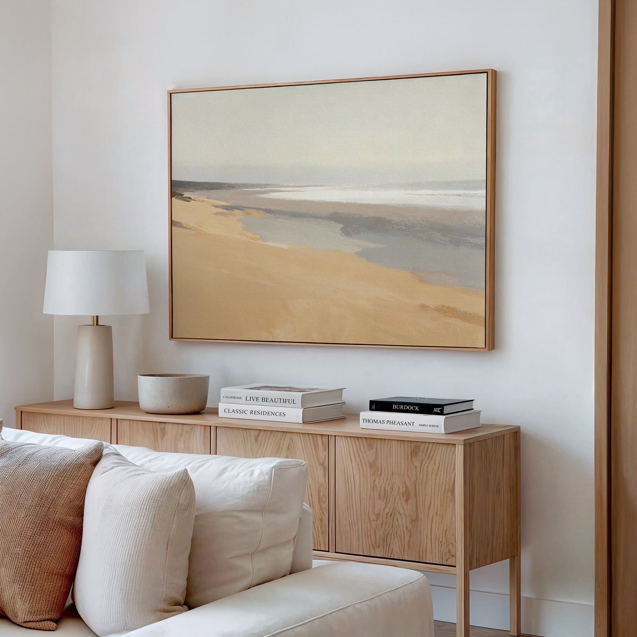 A minimalist living room featuring neutral furniture, a light wood sideboard with stacked books, a white lamp, a bowl, and the Tidal Meditation Canvas Art - Horizontal beach landscape hanging above.