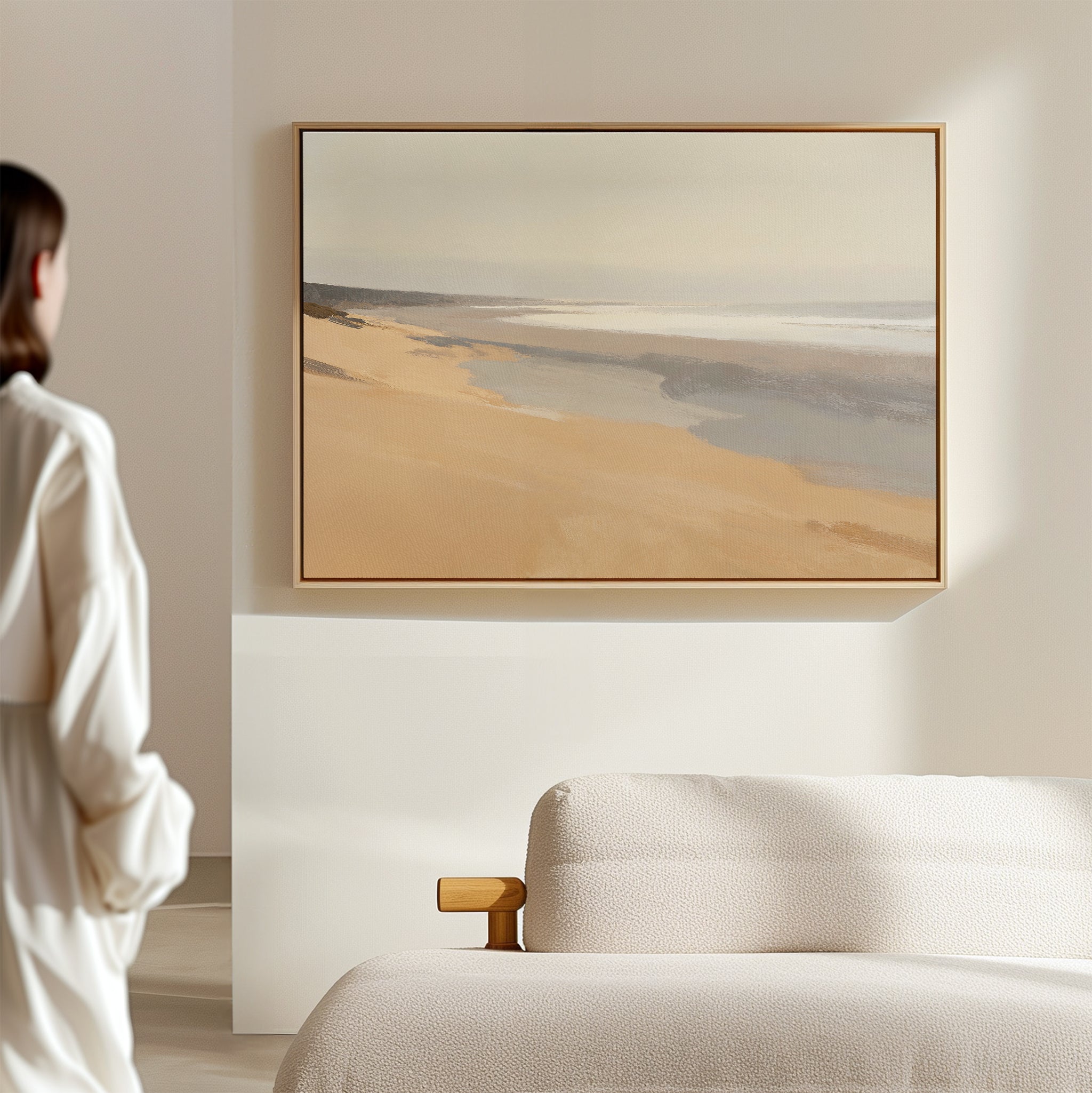 A woman in a white outfit stands in a minimal, sunlit room, admiring the Tidal Meditation Canvas Art - Horizontal, showcasing a serene beach scene above a textured beige sofa.