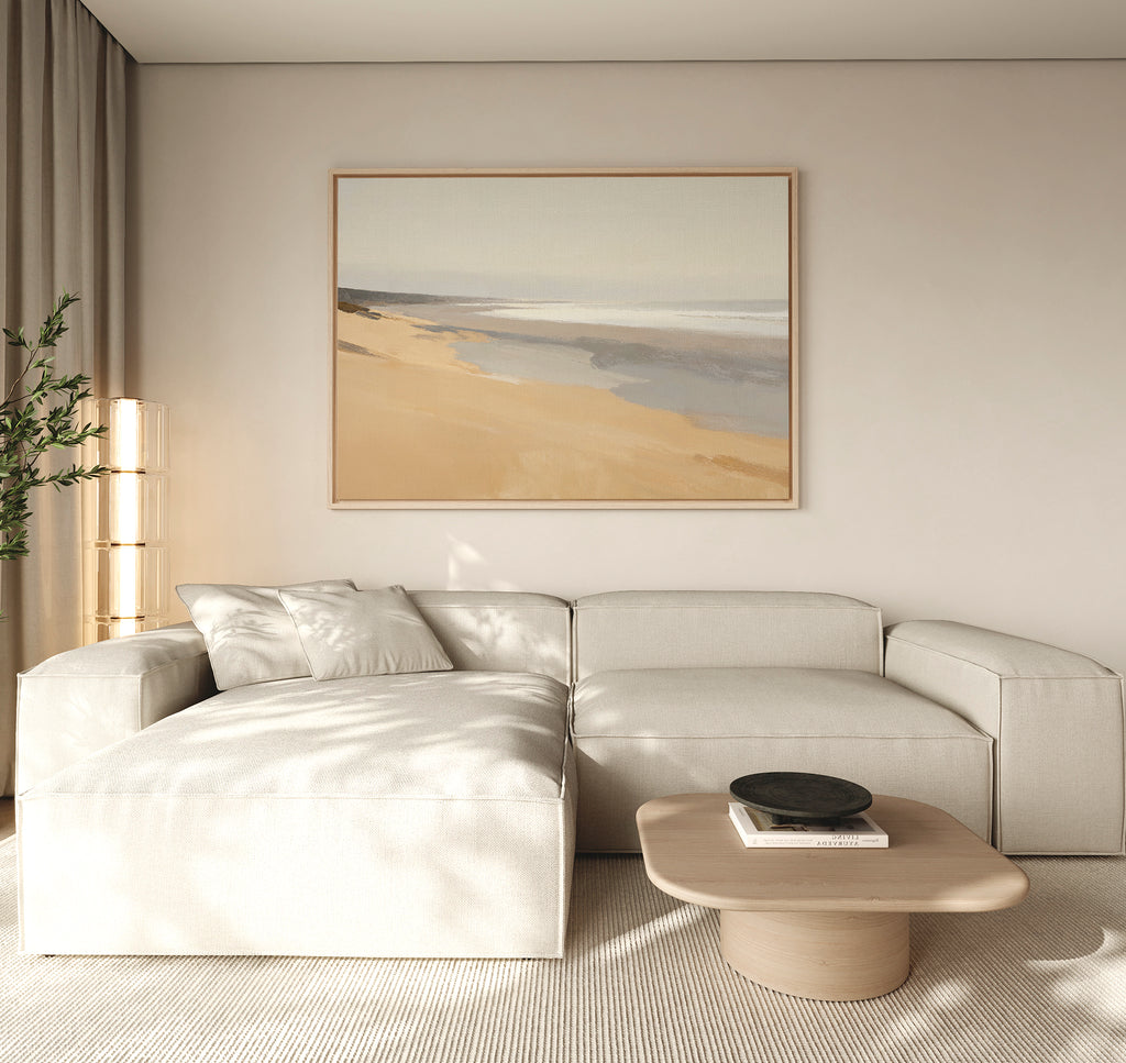 A minimal living room pairs a light beige sectional with a light wood coffee table. Above, the Tidal Meditation Canvas Art - Horizontal adds tranquility. Beige curtains let in natural light, and a leafy plant brings freshness to the corner.