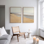 A modern living room features a rattan chair, small white table, lit candles, gray sofa, and the Tidal Meditation - Set of 2 canvas prints on a white wall near a large window for tranquil decor.
