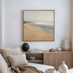 A cozy, neutral living room features a beige sofa, striped cushions, wooden decor, and the "Tidal Meditation Canvas Art - Square"—a tranquil beach scene—hung above a sideboard with stacked books and ceramics.