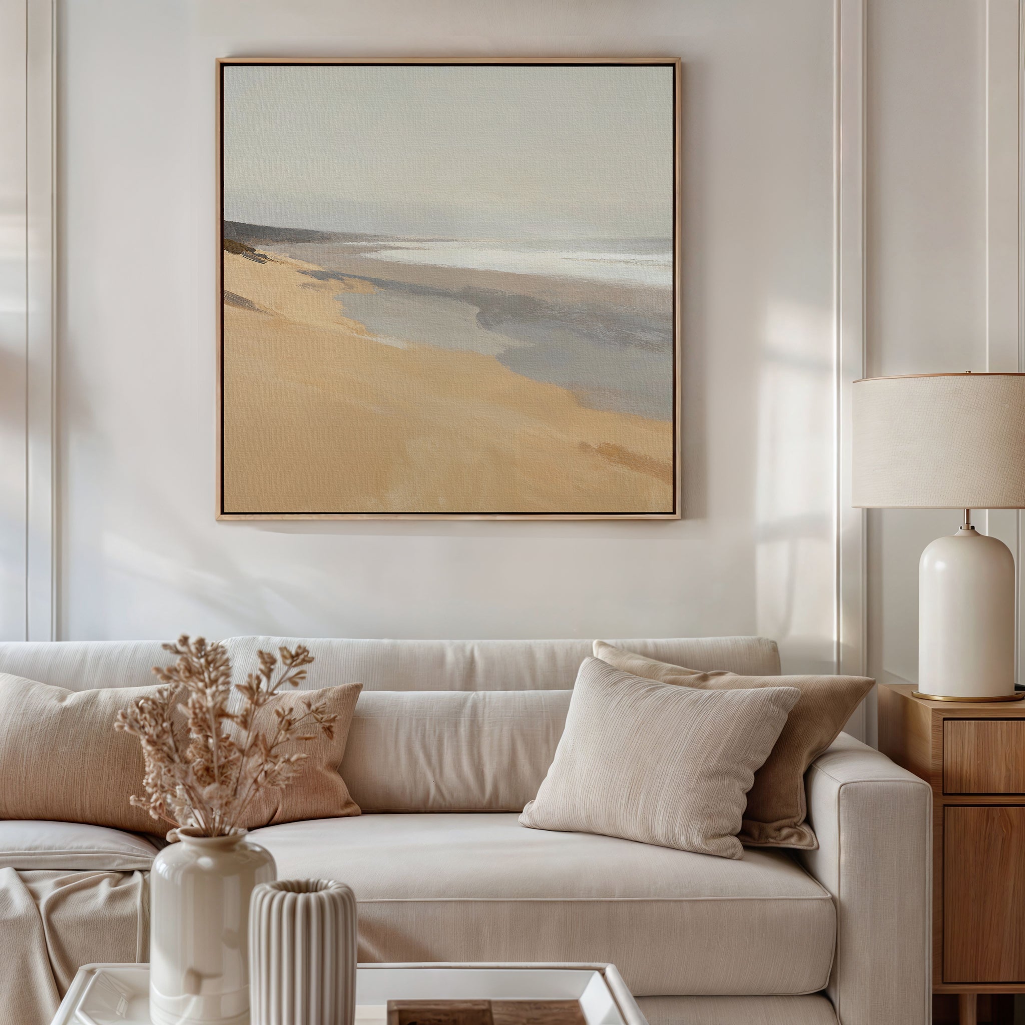 A cozy living room with a beige sofa, neutral pillows, a side table and lamp, dried flowers in a vase, and the Tidal Meditation Canvas Art - Square adding tranquil coastal vibes to the light-colored wall.