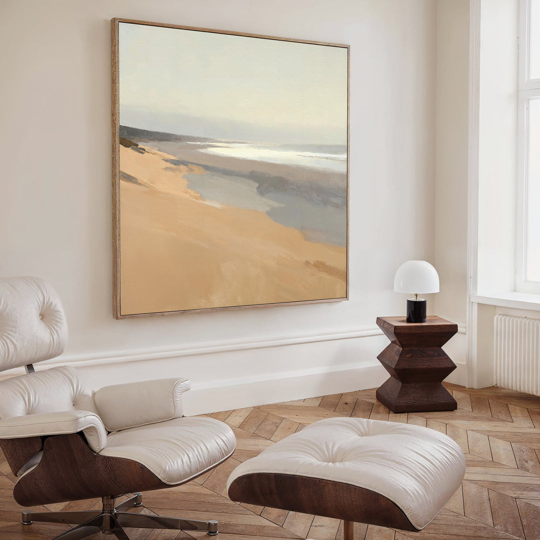 A modern living room with a white armchair and ottoman, a geometric wooden side table with a lamp, and the Tidal Meditation Canvas Art - Square, featuring a coastal scene, hanging on the light-colored wall.