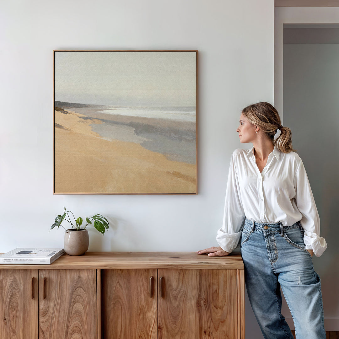 A woman in a white blouse and jeans stands by a wooden cabinet topped with a plant and book, gazing at the Tidal Meditation Canvas Art - Square displayed on the wall. The calm, modern setting highlights the tranquil coastal artwork.