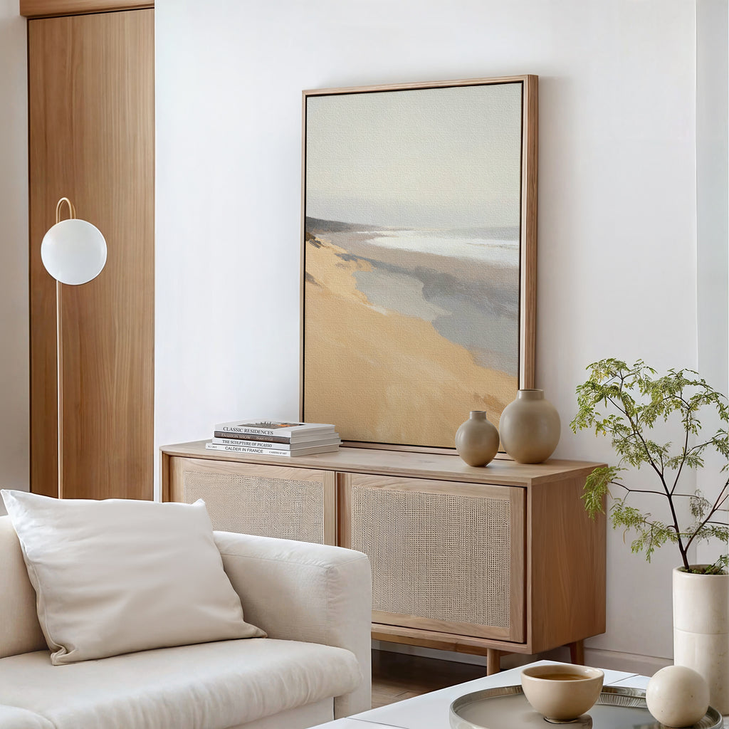 A modern living room with light wood furniture, a white sofa, beige vases, and a cushion is enhanced by the tranquil "Tidal Meditation Canvas Art - Vertical," bringing beach-inspired calm, while a small plant completes the minimalist space.