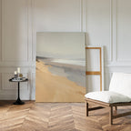 The Tidal Meditation Canvas Art - Vertical rests against a white paneled wall in a minimal room with herringbone wood floors, a cushioned chair, and a small black table holding candles and books.