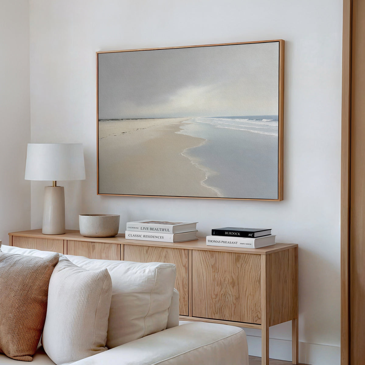 A modern living room features a wooden sideboard, white lamp, books, and neutral decor. Above hangs the Tidal Reflections Canvas Art - Horizontal—a large framed coastal print with soft colors for calming wall decor.