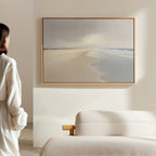 A person in a white robe stands by a beige sofa, admiring the Tidal Reflections Canvas Art - Horizontal, framed on a light wall in a modern minimalist room.