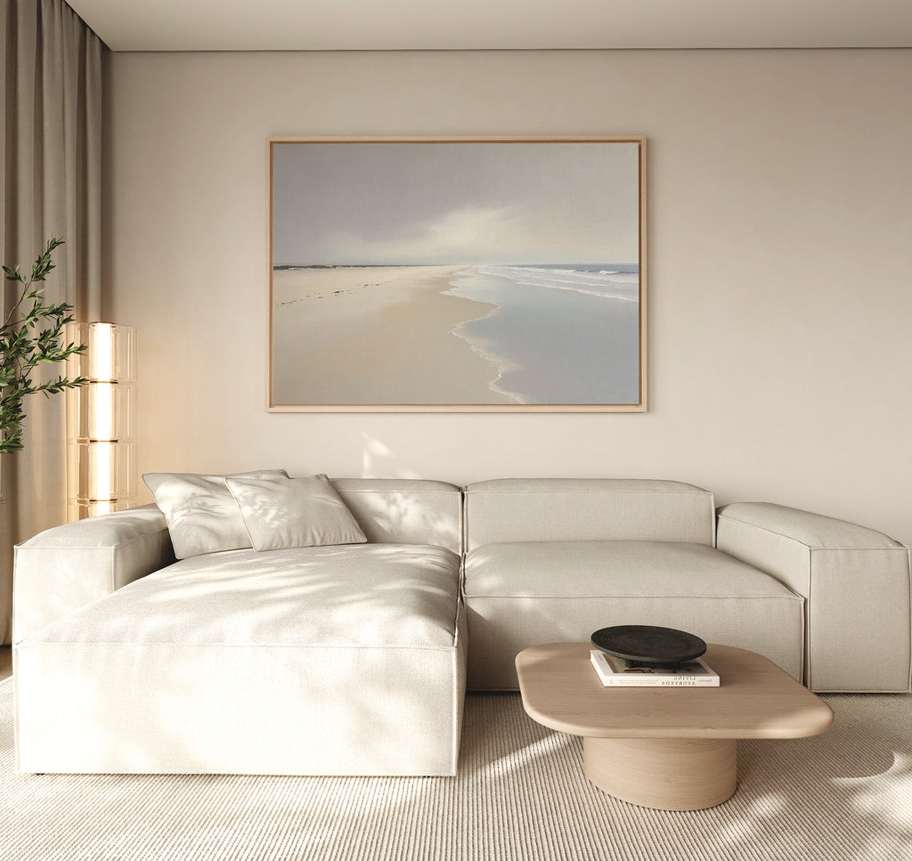 A minimalist living room features a beige sectional, light wood coffee table, potted plant, and the Tidal Reflections Canvas Art - Horizontal. Soft natural light highlights this serene ocean wall decor for a calm atmosphere.