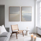A modern living room with a light gray sofa, wooden chair, small white side table with a lit candle, and Tidal Reflections - Set of 2 ocean art prints on the wall. Sunlight pours in through large windows.
