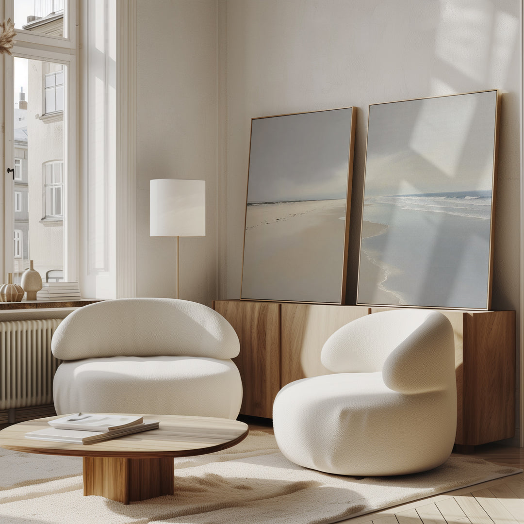Two modern, white, rounded armchairs and a low wooden table sit in a bright room with large windows. The Tidal Reflections - Set of 2 ocean art rests on a wooden sideboard, bringing a calm, airy feel to the space.