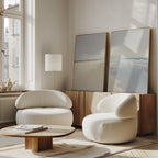 Two modern, white, rounded armchairs and a low wooden table sit in a bright room with large windows. The Tidal Reflections - Set of 2 ocean art rests on a wooden sideboard, bringing a calm, airy feel to the space.