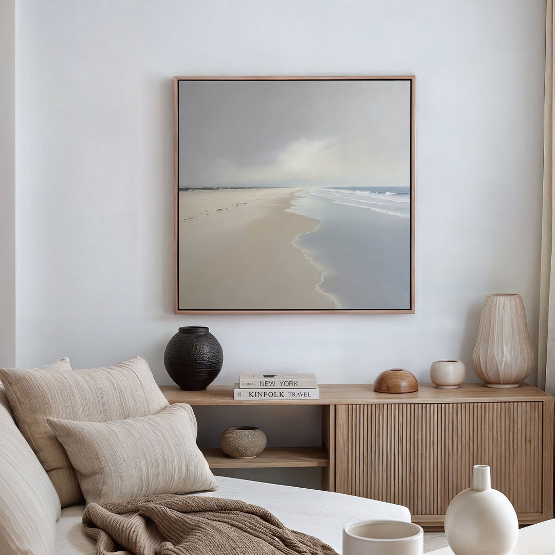 A serene, minimalist living room with a beige sofa, neutral cushions, wooden sideboard, decorative vases, and calming wall decor—a large Tidal Reflections Canvas Art - Square print of a calm beach hanging on a white wall.