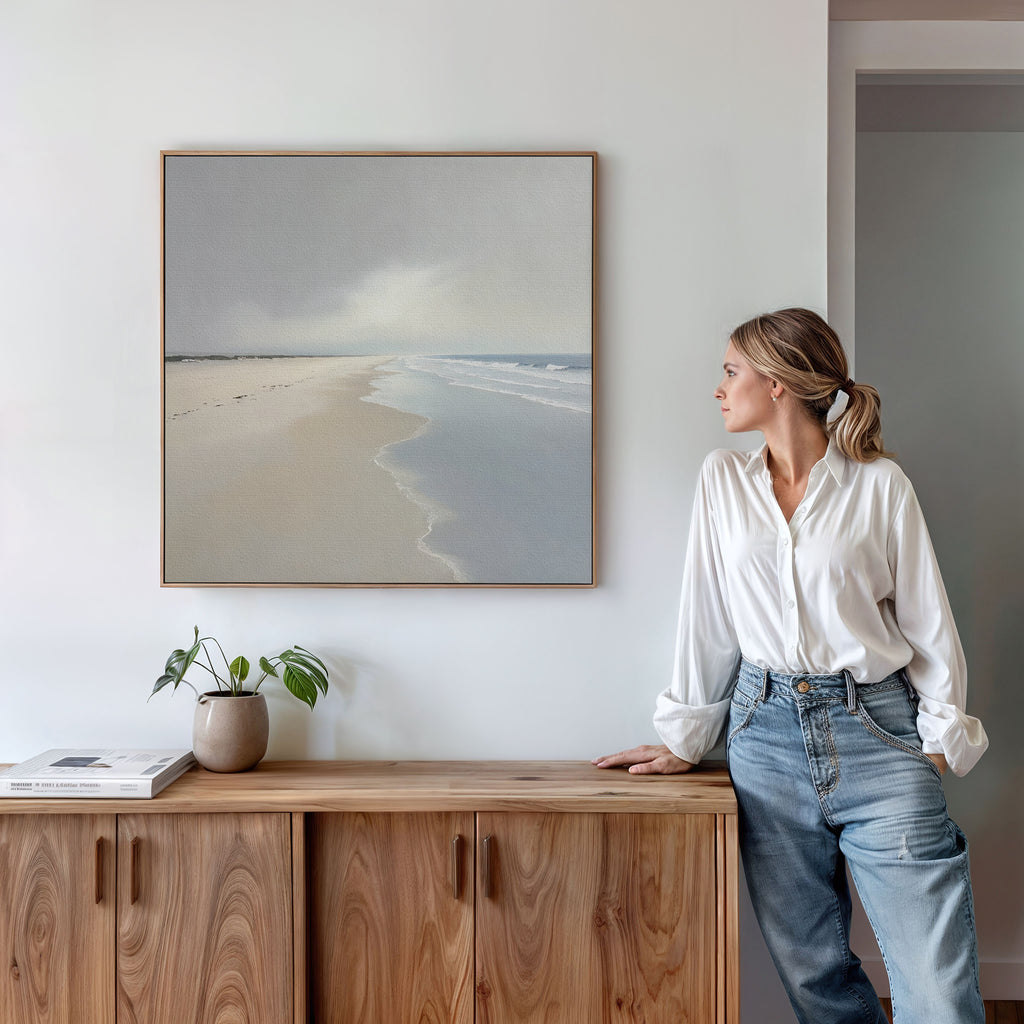 A woman in a white blouse and jeans leans on a wooden cabinet, gazing at the Tidal Reflections Canvas Art - Square hanging above. A plant and books on the cabinet enhance the calming decor of the serene space.