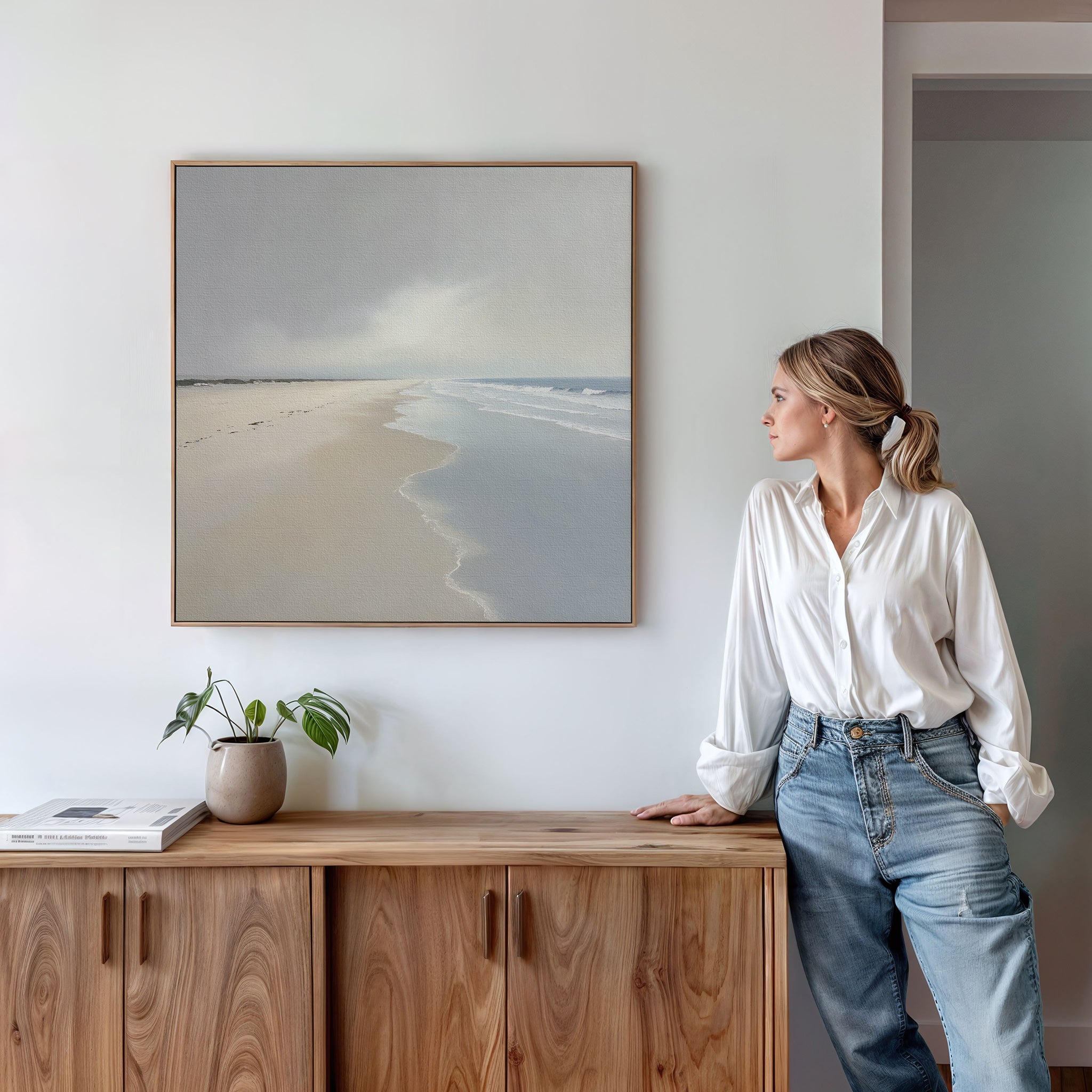 A woman in a white blouse and jeans leans on a wooden cabinet, gazing at the Tidal Reflections Canvas Art - Square hanging above. A plant and books on the cabinet enhance the calming decor of the serene space.