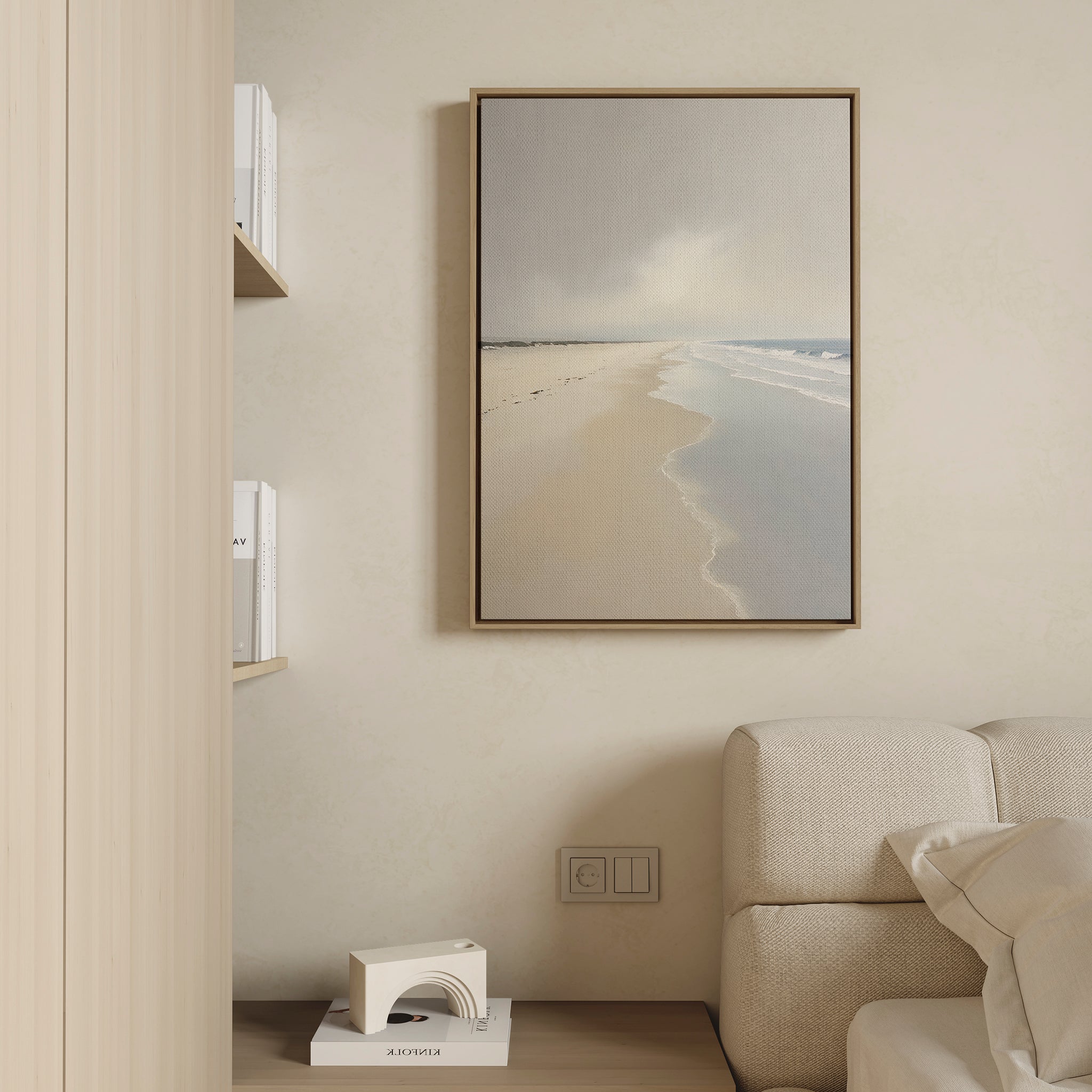The Tidal Reflections Canvas Art - Vertical, featuring a serene beach scene, is displayed in a frame above a light sofa on a beige wall. On adjacent shelves, white books and a geometric white decor accent are neatly arranged.