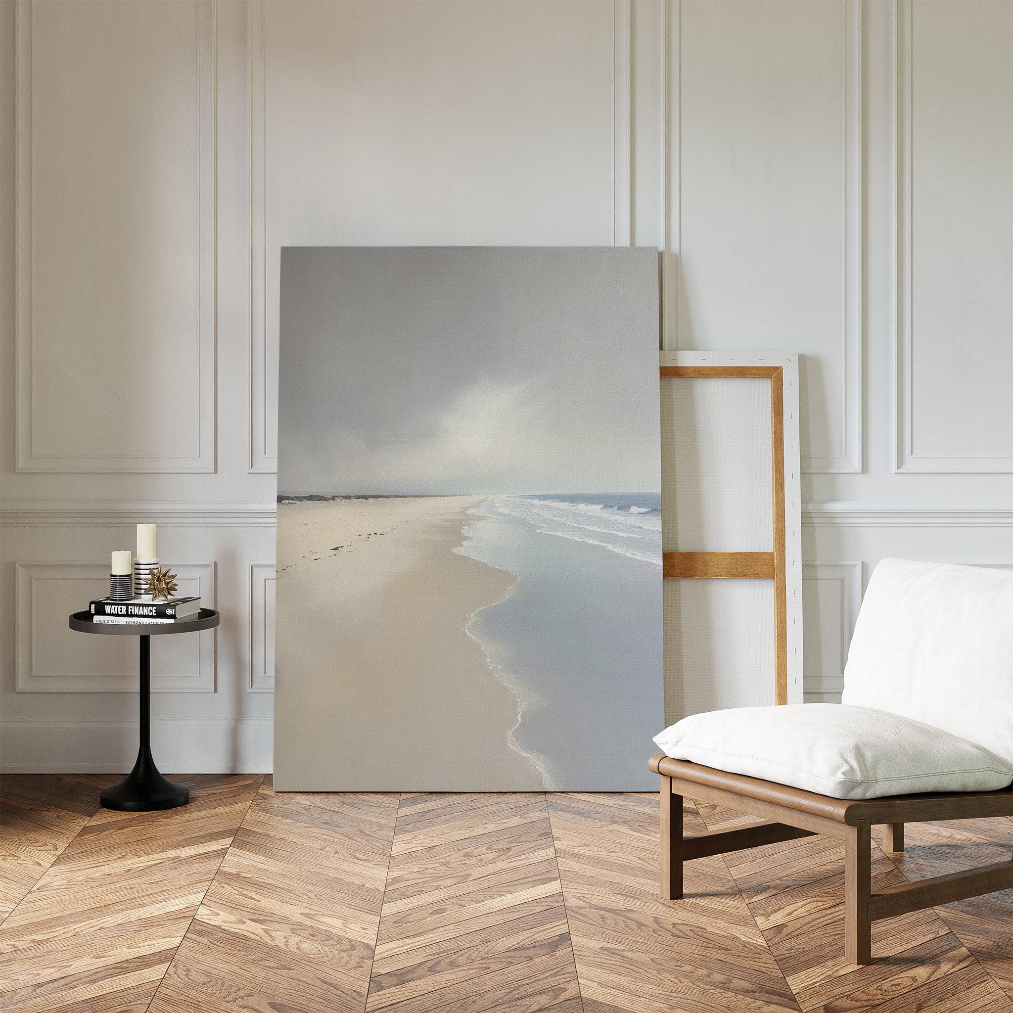 The Tidal Reflections Canvas Art - Vertical, featuring a serene beach scene, leans against a white paneled wall beside a wooden chair with a white cushion and a black side table holding candles and a book on the herringbone floor.